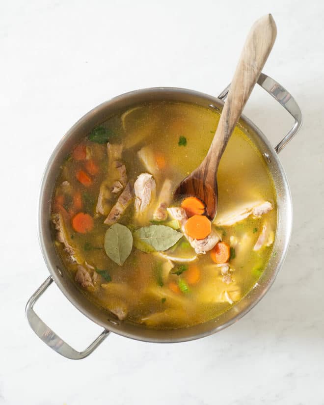 a large stainless steel pot of chicken noodle soup (before the noodles have been added in)with a wooden spoon sticking out of it