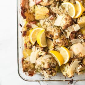 a casserole dish of chicken thigh pesto bake that just came out of the oven and is done baking, garnished with lemon wedges, parmesan cheese, and pine nuts