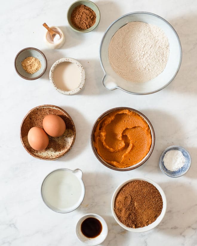 all of the ingredients for healthy pumpkin bread in different sized bowls on a marble surface