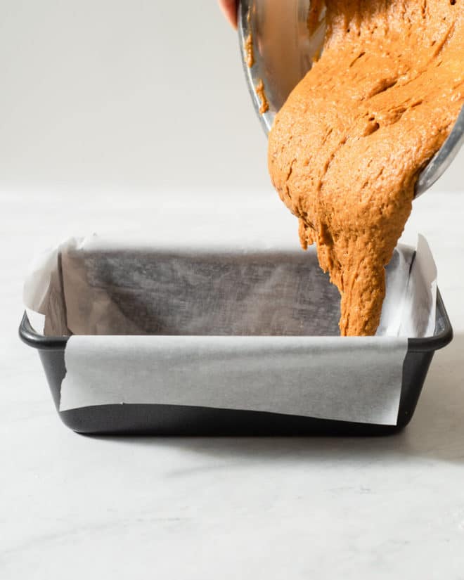 a person pouring vegan pumpkin bread batter into a parchment paper lined loaf pan