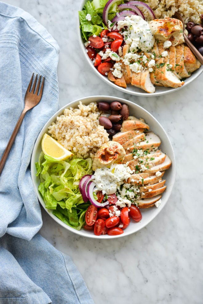 a Greek chicken power bowl filled with lettuce, tomatoes, chicken, olives, brown rice, hummus, tzatziki, onions, and feta