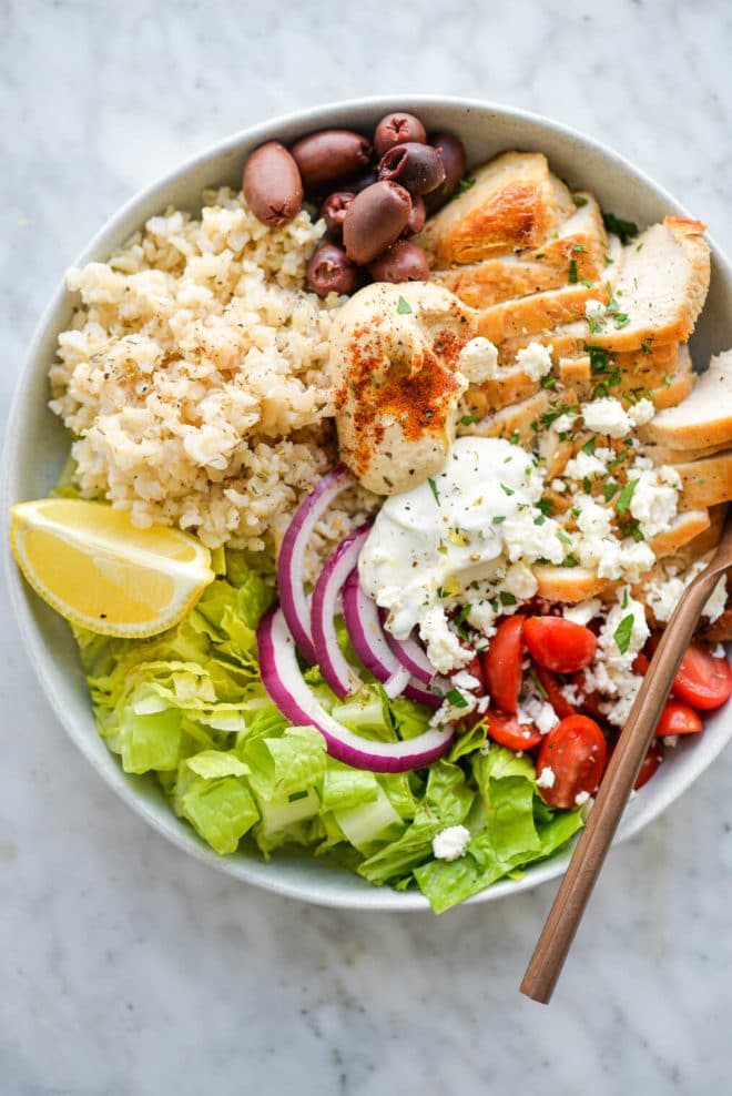 a green chicken power bowl made with brown rice, chicken, olives, cherry tomatoes, hummus, and lettuce