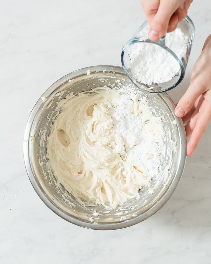 a person adding powdered sugar into a metal bowl of cream cheese frosting
