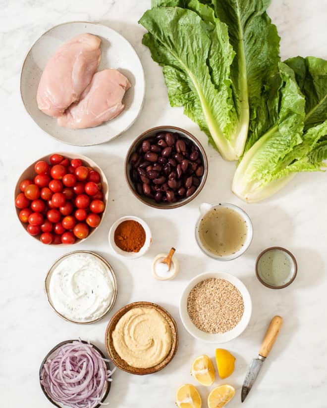 all of the ingredients for Greek chicken bowls in separate bowls and containers on a marble surface