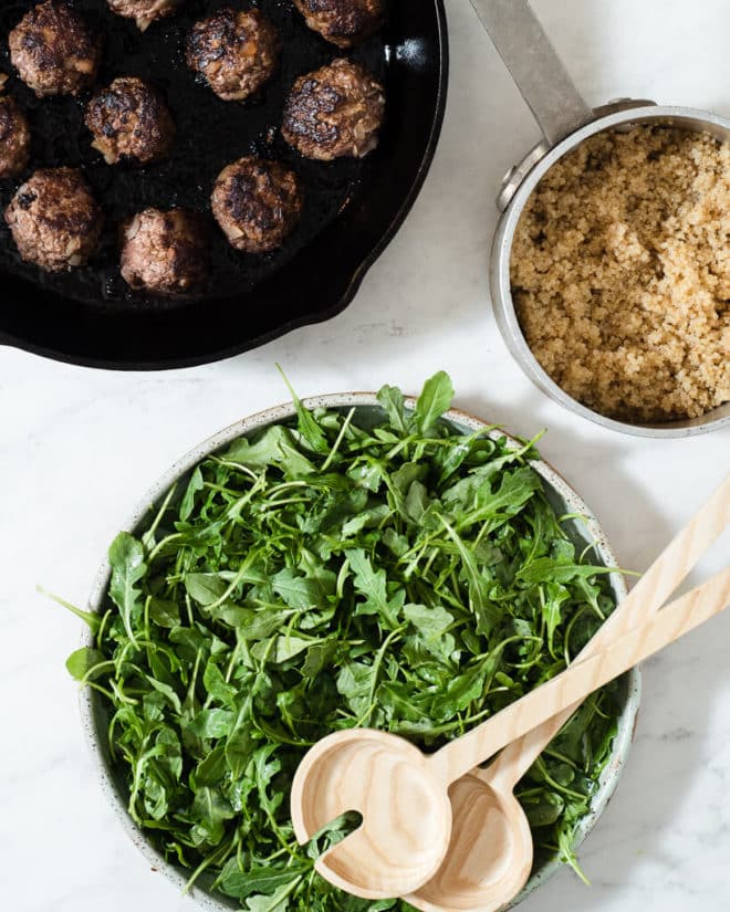 a cast iron skillet of greek meatballs, a pot of white quinoa, and a bowl of simple arugula salad on a marble surface