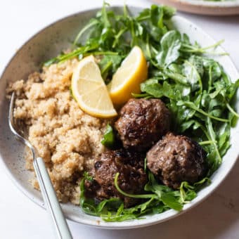 a serving of greek meatballs, arugula salad, and white quinoa served with 2 lemon wedges