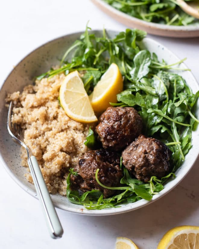 a serving of greek meatballs, arugula salad, and white quinoa served with 2 lemon wedges