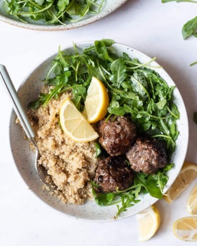 a serving of greek meatballs, arugula salad, and white quinoa served with 2 lemon wedges
