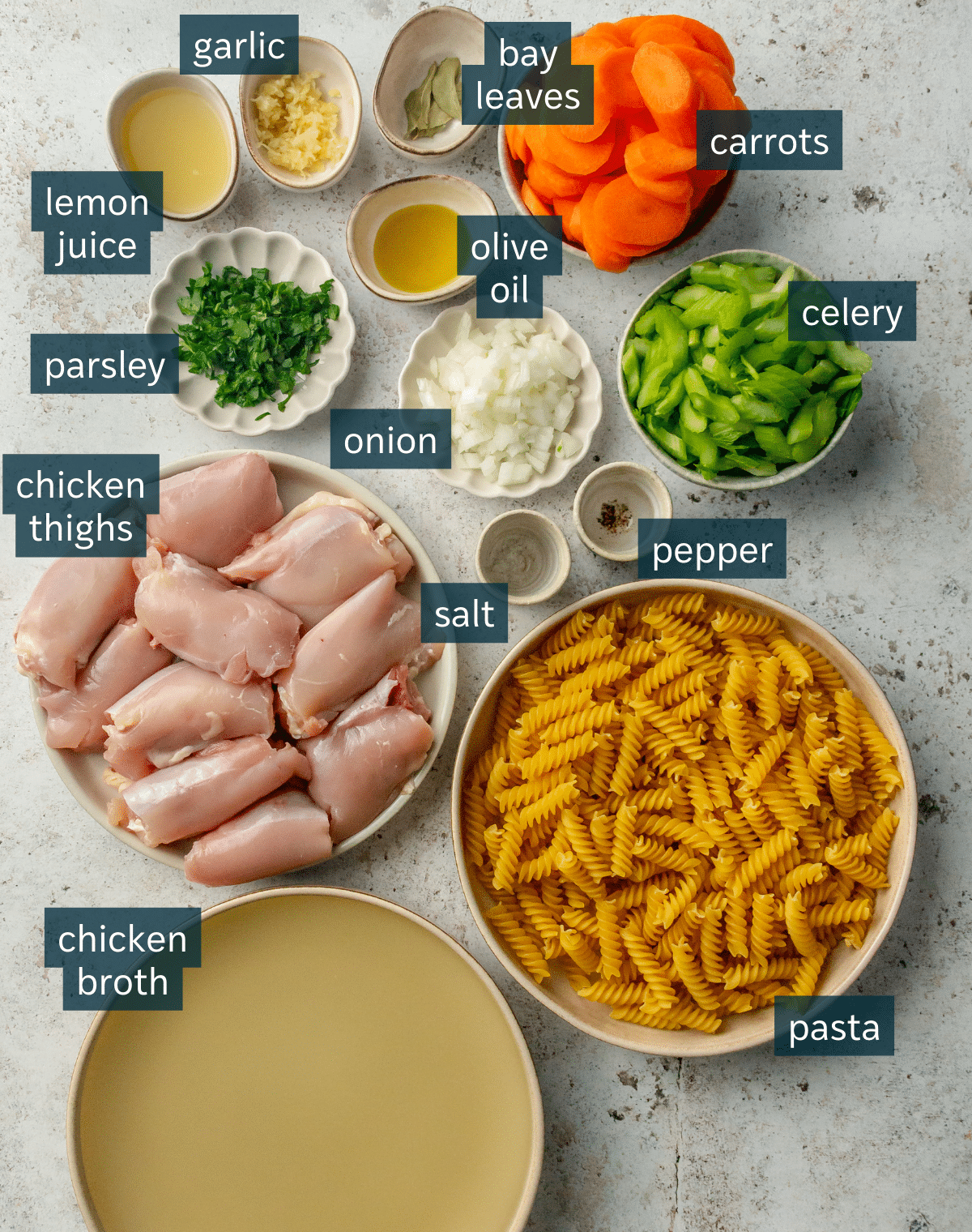 All of the ingredients for homemade chicken noodle soup in different sized bowls and plates on a light gray surface.
