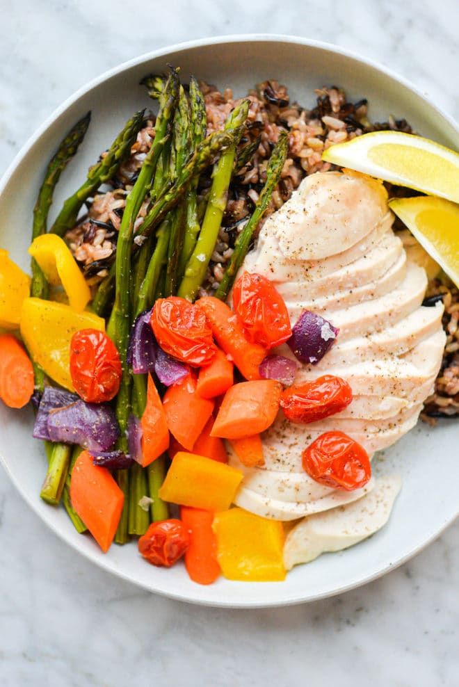 a plate of roasted chicken breast, roasted veggies, and wild rice served with two lemon wedges