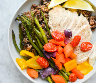 a plate of roasted chicken breast, roasted veggies, and wild rice served with two lemon wedges