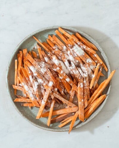 a person sprinkling arrowroot starch over a plate of cut sweet potato fries