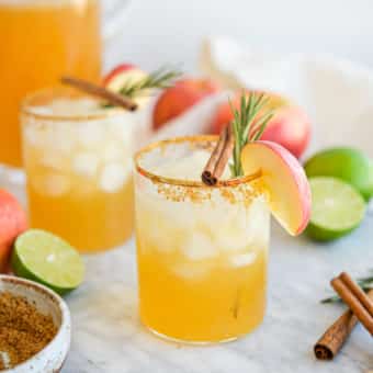 two apple cider margaritas next to a pitcher of margaritas next to apples, limes, brown sugar, cinnamon sticks, and rosemary