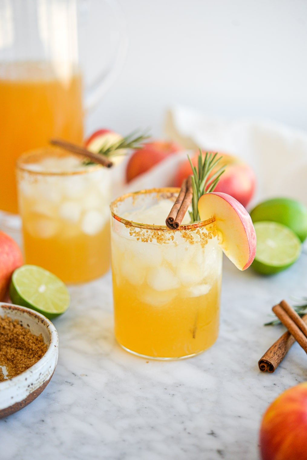 two apple cider margaritas next to a pitcher of margaritas next to apples, limes, brown sugar, cinnamon sticks, and rosemary