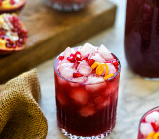 two pomegranate margaritas sitting in front of a bowl of pomegranate arils and whole pomegranates