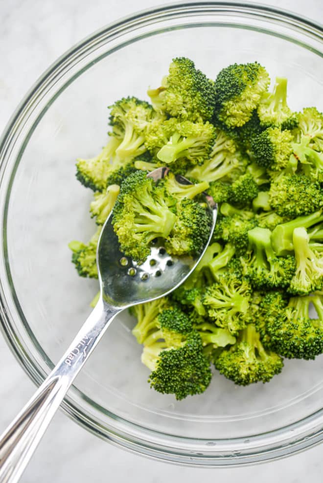 a glass bowl of steamed broccoli with a spoonful of it being held over the bowl