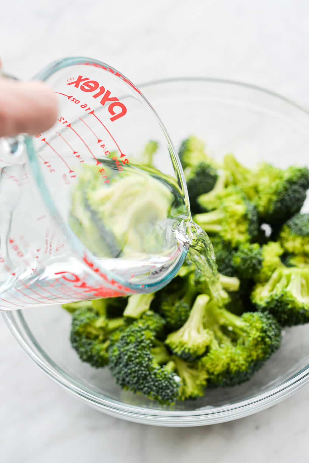 a person pouring water into a glass bowl of broccoli florets