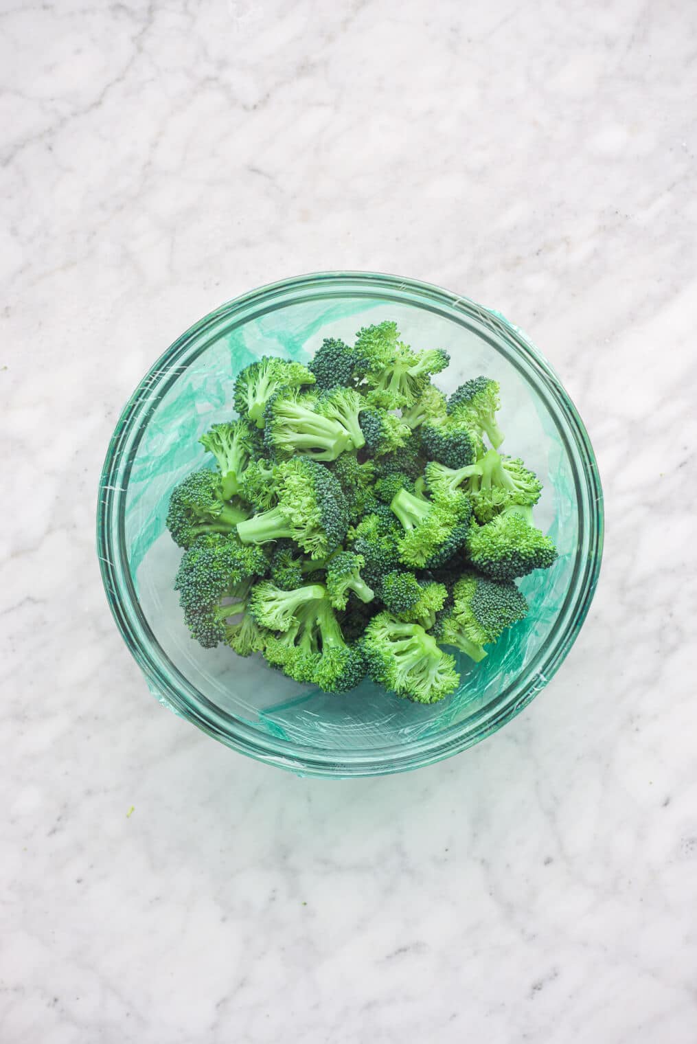 a glass bowl of steamed broccoli covered in saran wrap