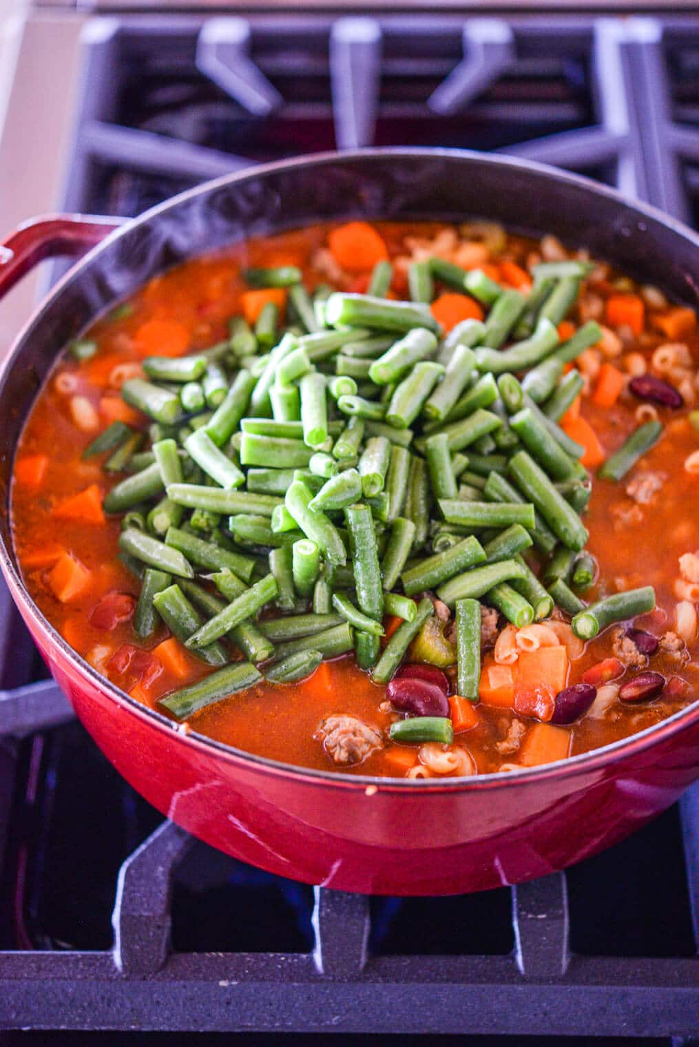 green beans being added to a large pot of minestrone soup on the stovetop