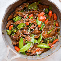 Cooked snow peas, sliced onion, sliced carrot, broccoli florets, and sliced beef with brown sauce topped with sesame seeds in grey sauté pan with sand colored handle