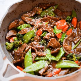 Cooked snow peas, sliced onion, sliced carrot, broccoli florets, and sliced beef with brown sauce topped with sesame seeds in grey sauté pan with sand colored handle