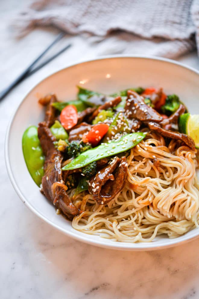 Bowl of beef stir fry containing noodles, beef, snow peas, sliced carrots, broccoli, and wedge of lime sprinkled with sesame seeds on white and grey marble countertop