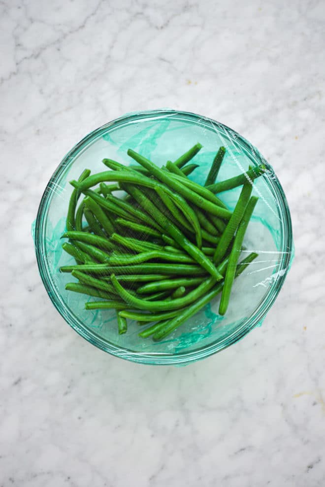 Glass bowl with green string beans covered with green plastic wrap sitting on grey and white marble countertop