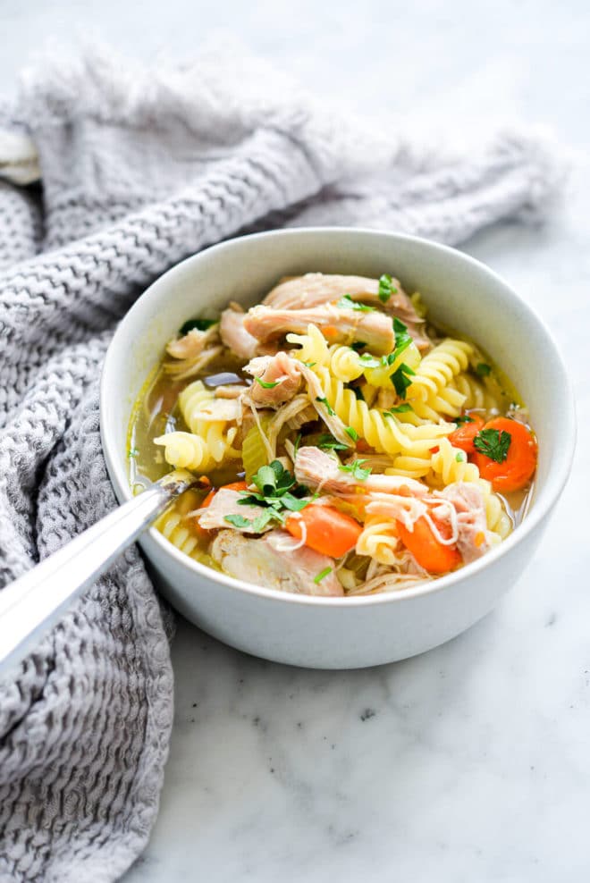 Bowl of chicken noodle soup containing spiral noodles, shredded chicken, sliced carrots, and sprinkle of parsley with silver spoon wrapped with grey, textured hand towel on left side and sitting on white and grey marble countertop