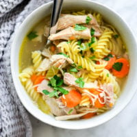Bowl of chicken noodle soup containing spiral noodles, shredded chicken, sliced carrots, and sprinkle of parsley with silver spoon wrapped with grey, textured hand towel on left side and sitting on white and grey marble countertop