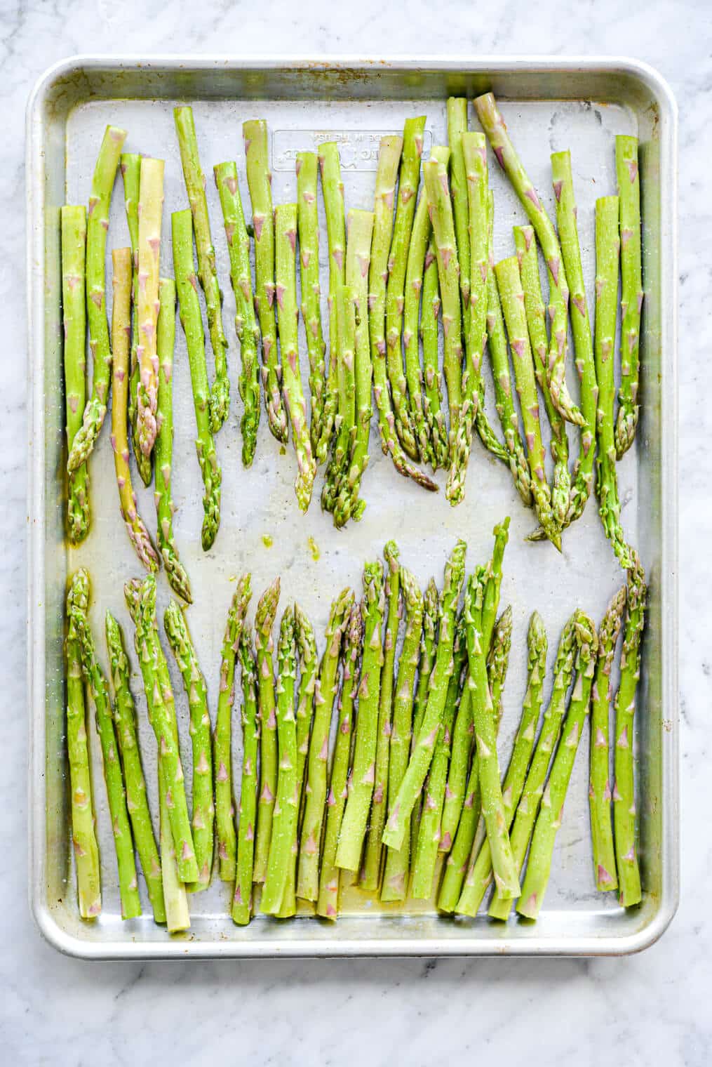 Aluminum sheet pan on grey and white marble countertop with two rows of asparagus