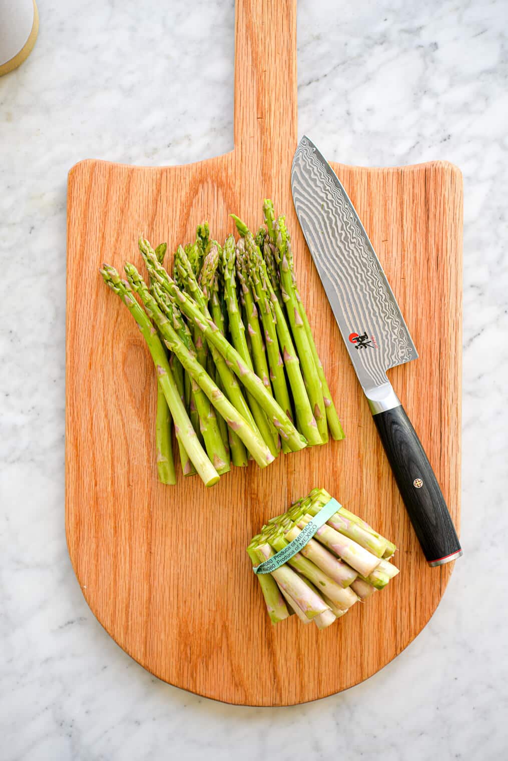 One bunch of asparagus with the bottoms chopped off just above the green rubber band sitting on a cutting board with a silver chef's knife with black handle