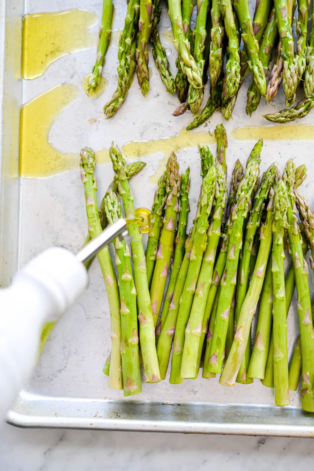 Metal sheet pan with two rows of asparagus being drizzled with olive oil from olive oil dispenser