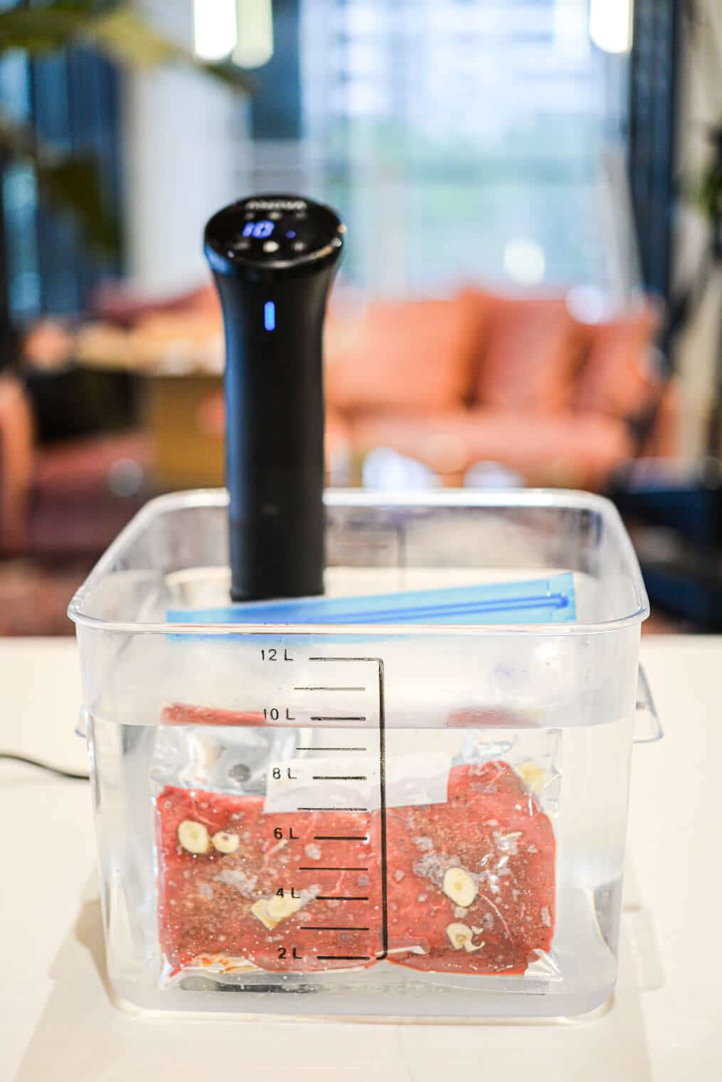 Large plastic container filled with water and black handled sous vide machine sitting on countertop with steaks cooking sous vide style inside