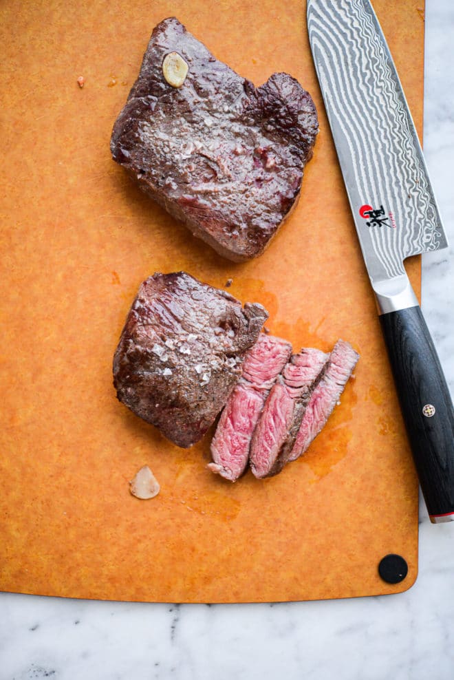 Two cooked steaks and chef's knife with black handle sitting on top of cutting board with three medium rare slices cut from one steak
