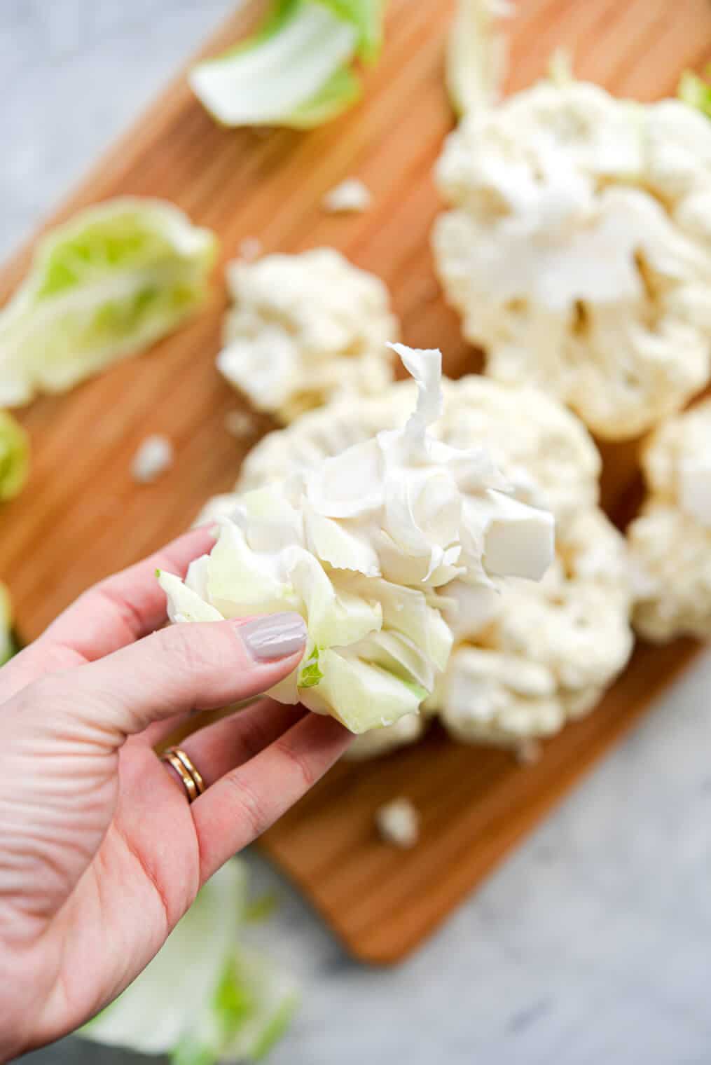 Close up of hand holding cauliflower stem with cauliflower florets and cutting board blurred in background