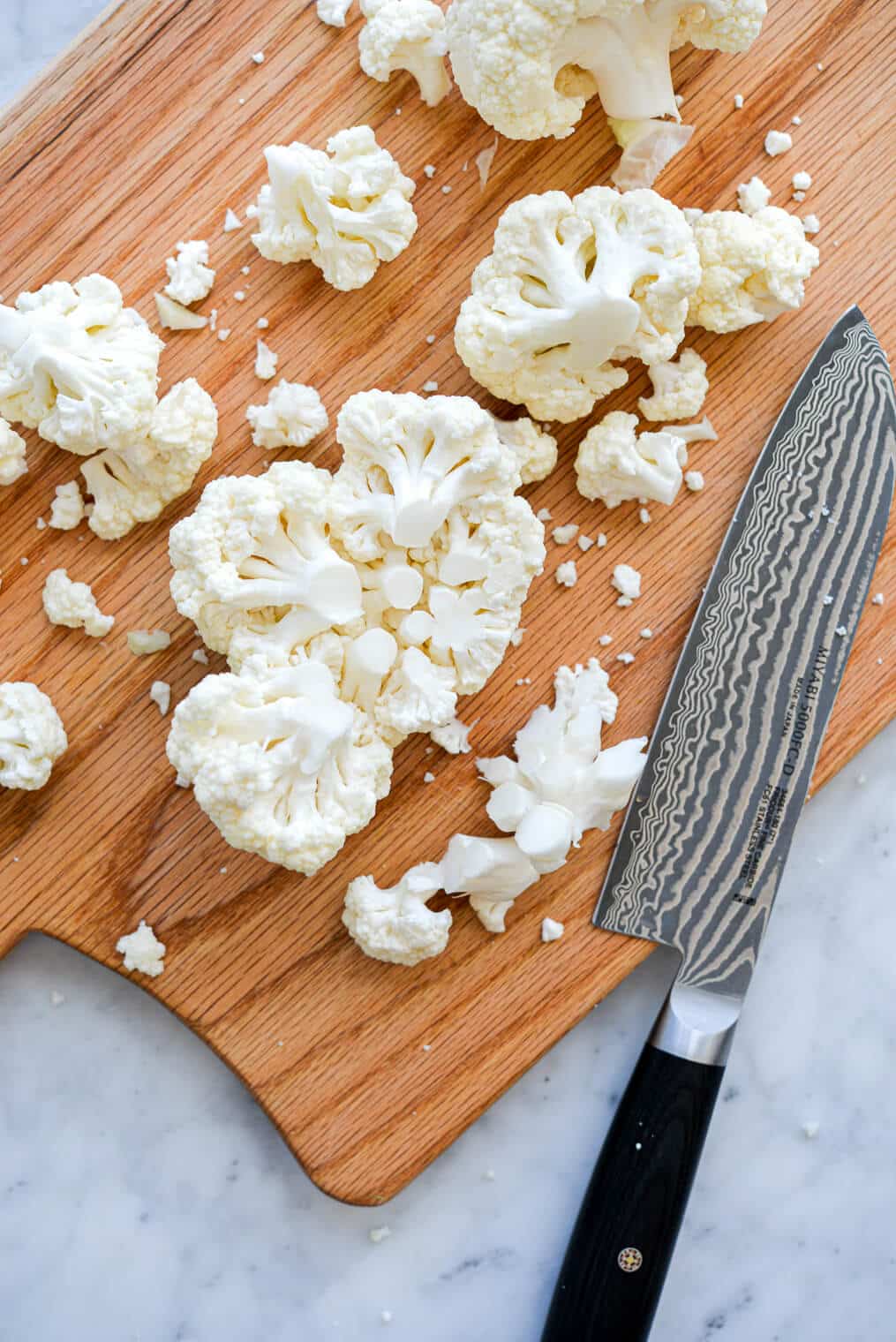 Wooden cutting board with cut cauliflower florets and chef's know with black handle sitting on grey and white marble countertop