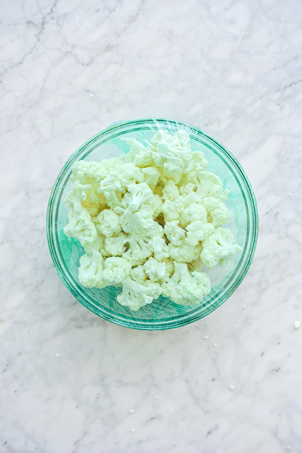 Cauliflower florets in glass bowl covered with light green plastic wrap sitting on grey and white marble countertop