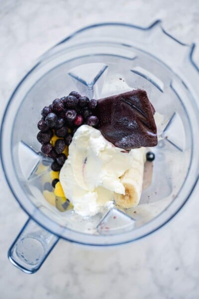 Top down view of blender with frozen blueberries, açaí puree, greek yogurt, sliced banana, and sliced mango