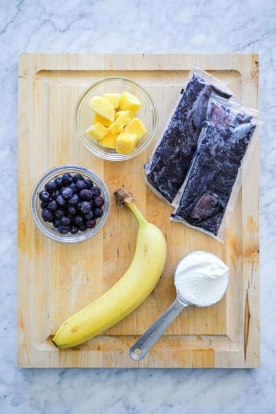Wooden cutting board with two packs of frozen açaí puree, bowl of frozen mango chunks, bowl of frozen blueberries, banana, and measuring cup with greek yogurt