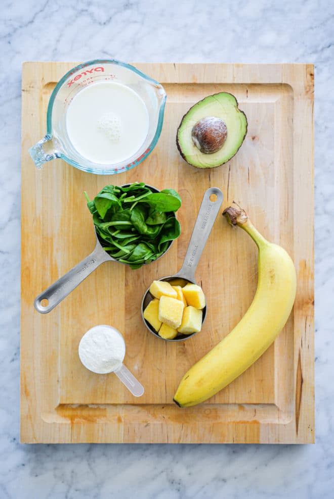Cutting board with milk in measuring glass, half an avocado, spinach in measuring cup, mango chunks in measuring cup, protein powder in measuring cup, and whole banana