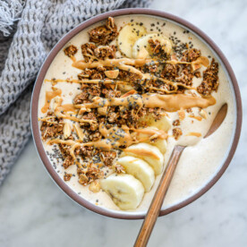 Banana smoothie bowl with spoon in bowl topped with sliced banana, granola, chia seeds, and a drizzle of peanut butter