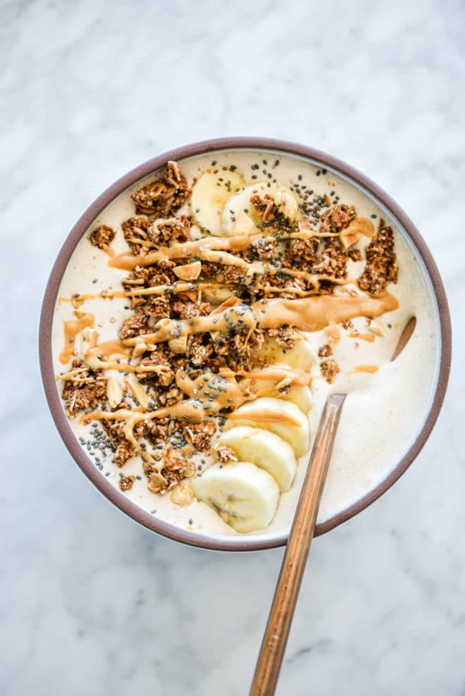 Banana smoothie bowl with spoon in bowl topped with sliced banana, granola, chia seeds, and a drizzle of peanut butter