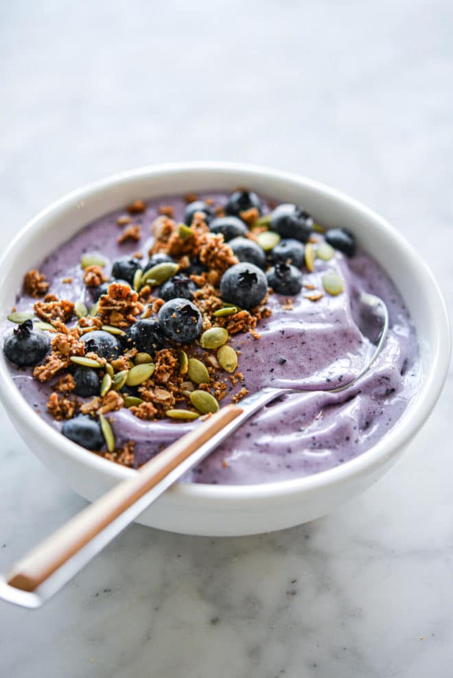 Blueberry smoothie with wooden and silver spoon in bowl topped with blueberries, pumpkin seeds, and granola