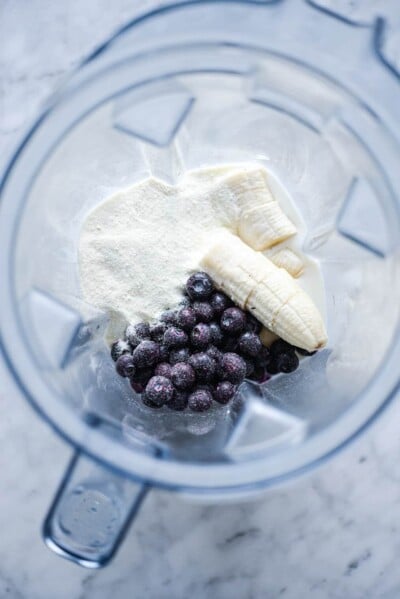 Top down view of blender with milk, protein powder, frozen blueberries, and sliced banana