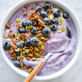 Blueberry smoothie with wooden and silver spoon in bowl topped with blueberries, pumpkin seeds, and granola