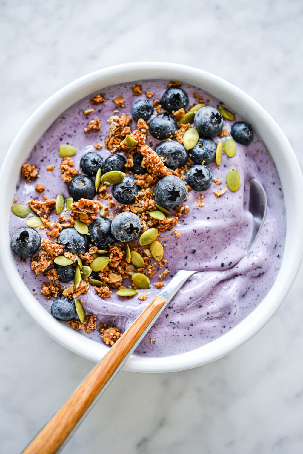 Blueberry smoothie with wooden and silver spoon in bowl topped with blueberries, pumpkin seeds, and granola