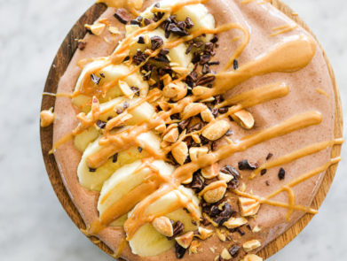 Chocolate smoothie in bowl topped with cocoa nibs, chopped peanuts, sliced banana, and a drizzle of peanut butter
