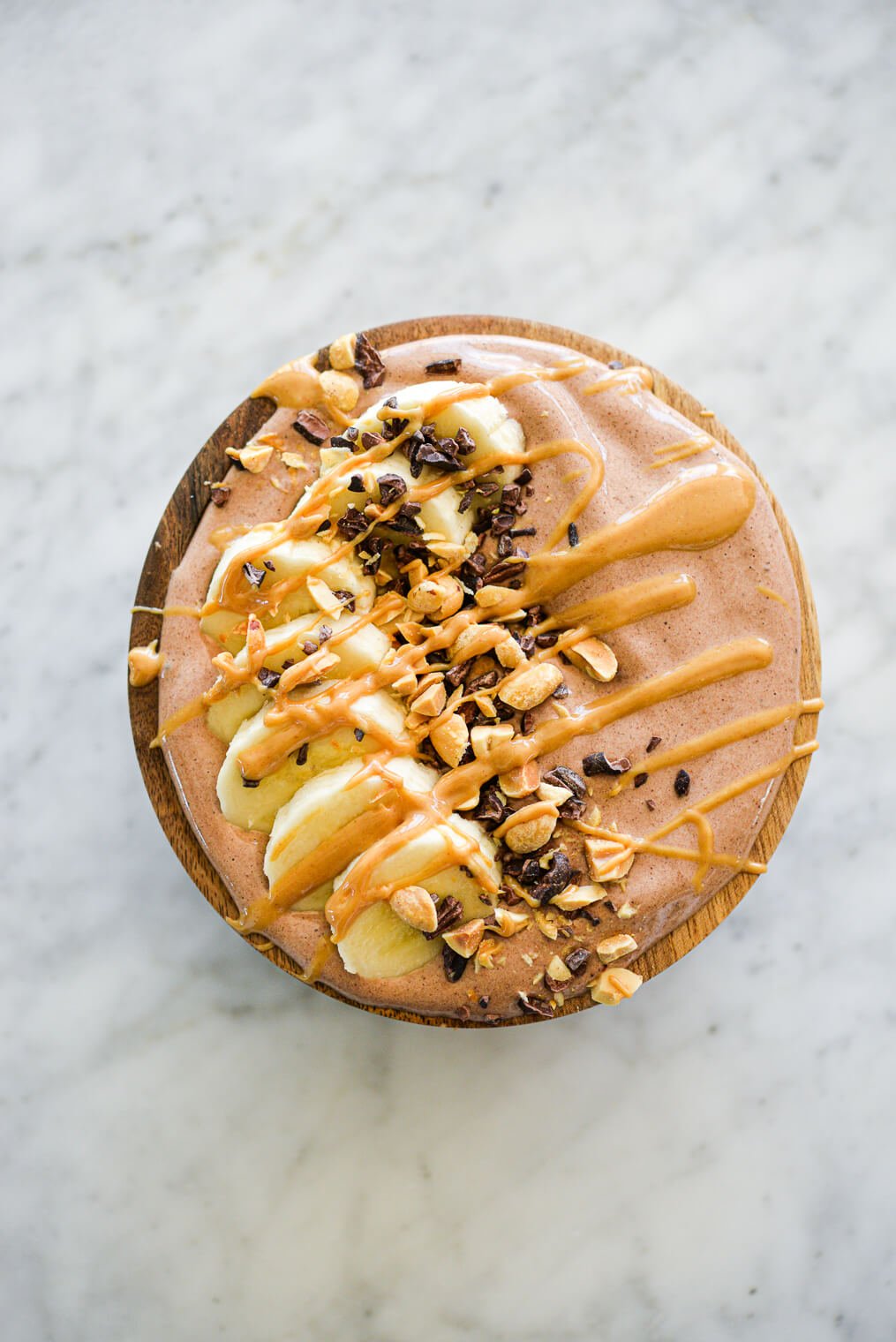 Chocolate smoothie in bowl topped with cocoa nibs, chopped peanuts, sliced banana, and a drizzle of peanut butter