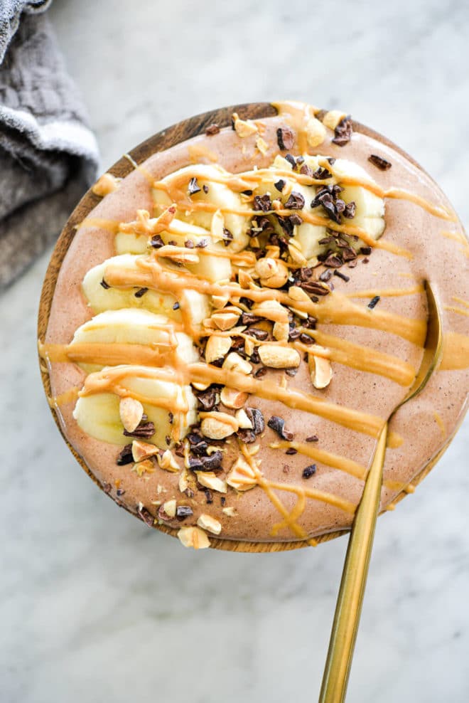 Chocolate smoothie with gold spoon in bowl topped with cocoa nibs, chopped peanuts, sliced banana, and a drizzle of peanut butter