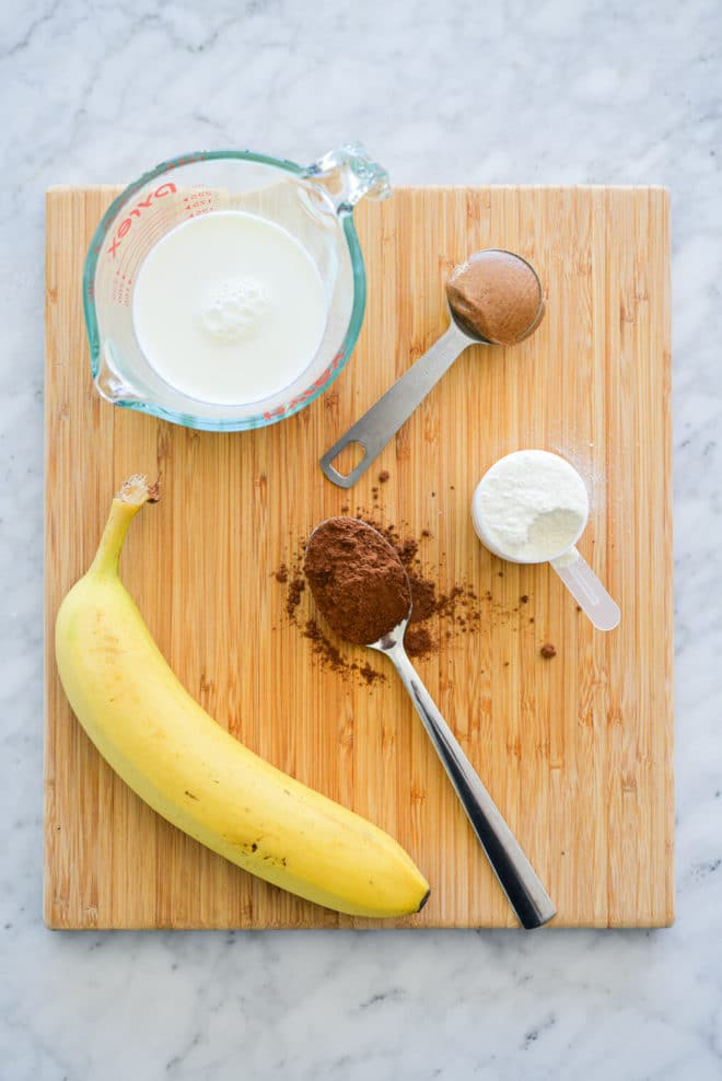 Cutting board with milk in measuring cup, peanut butter in measuring spoon, protein powder in measuring cup, cocoa powder on a spoon, and a whole banana